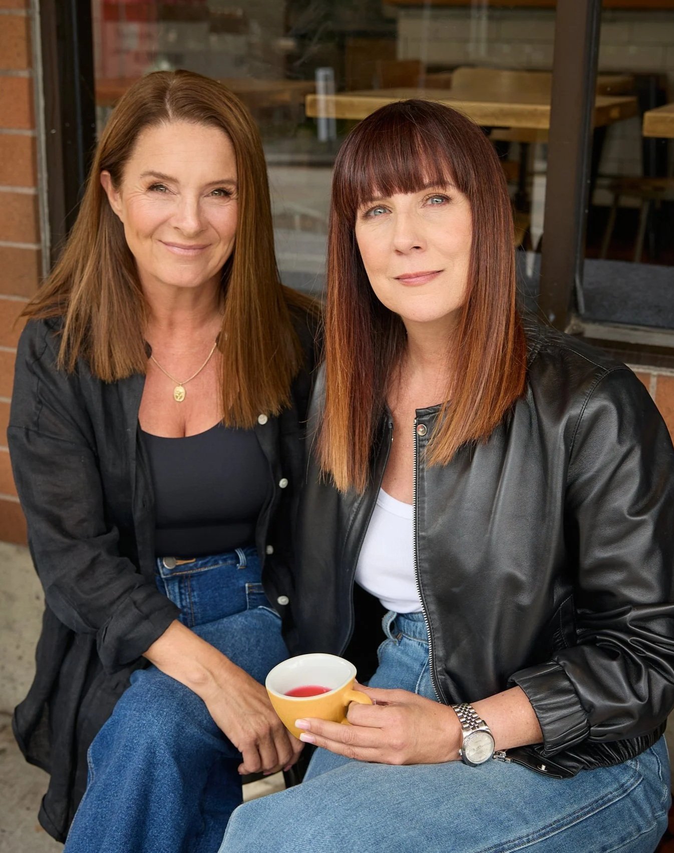 Two women with reddish-brown hair sitting outside a cafe, one holding a yellow cup, both smiling at the camera.