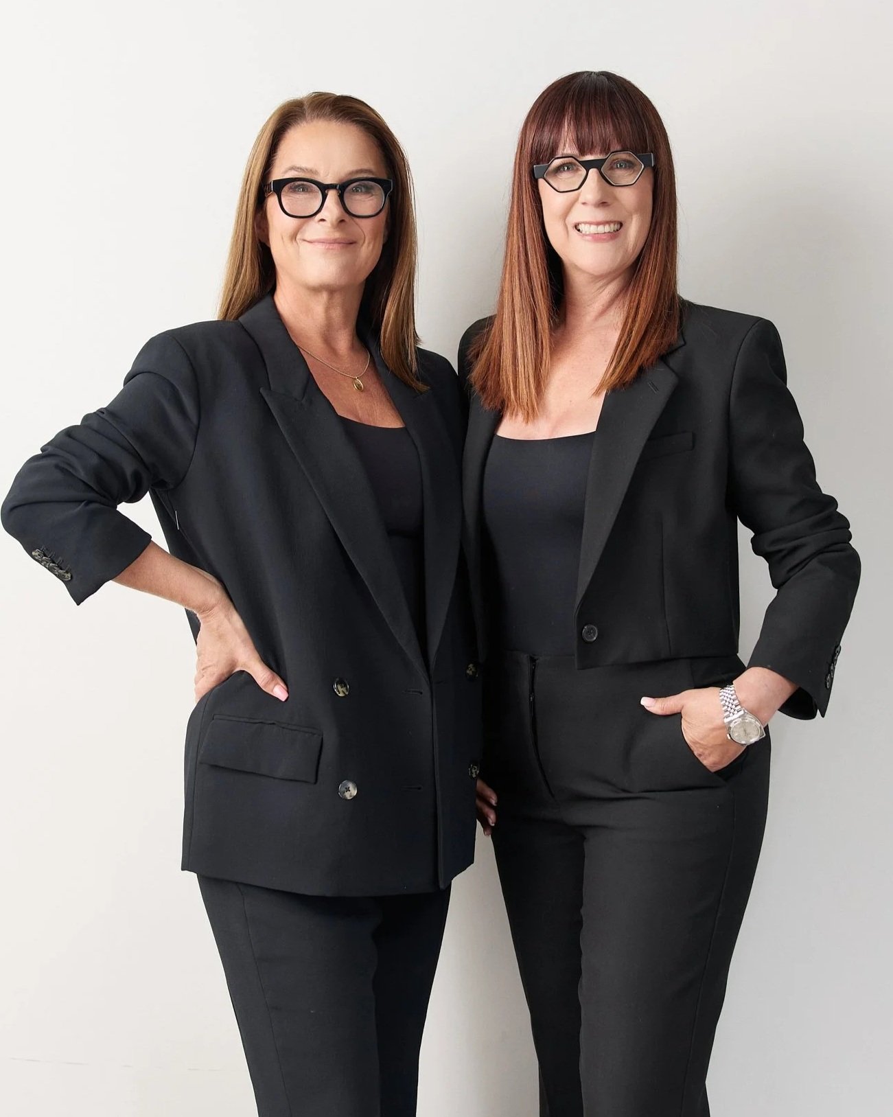 Two women in black suits and glasses posing against a plain white background.