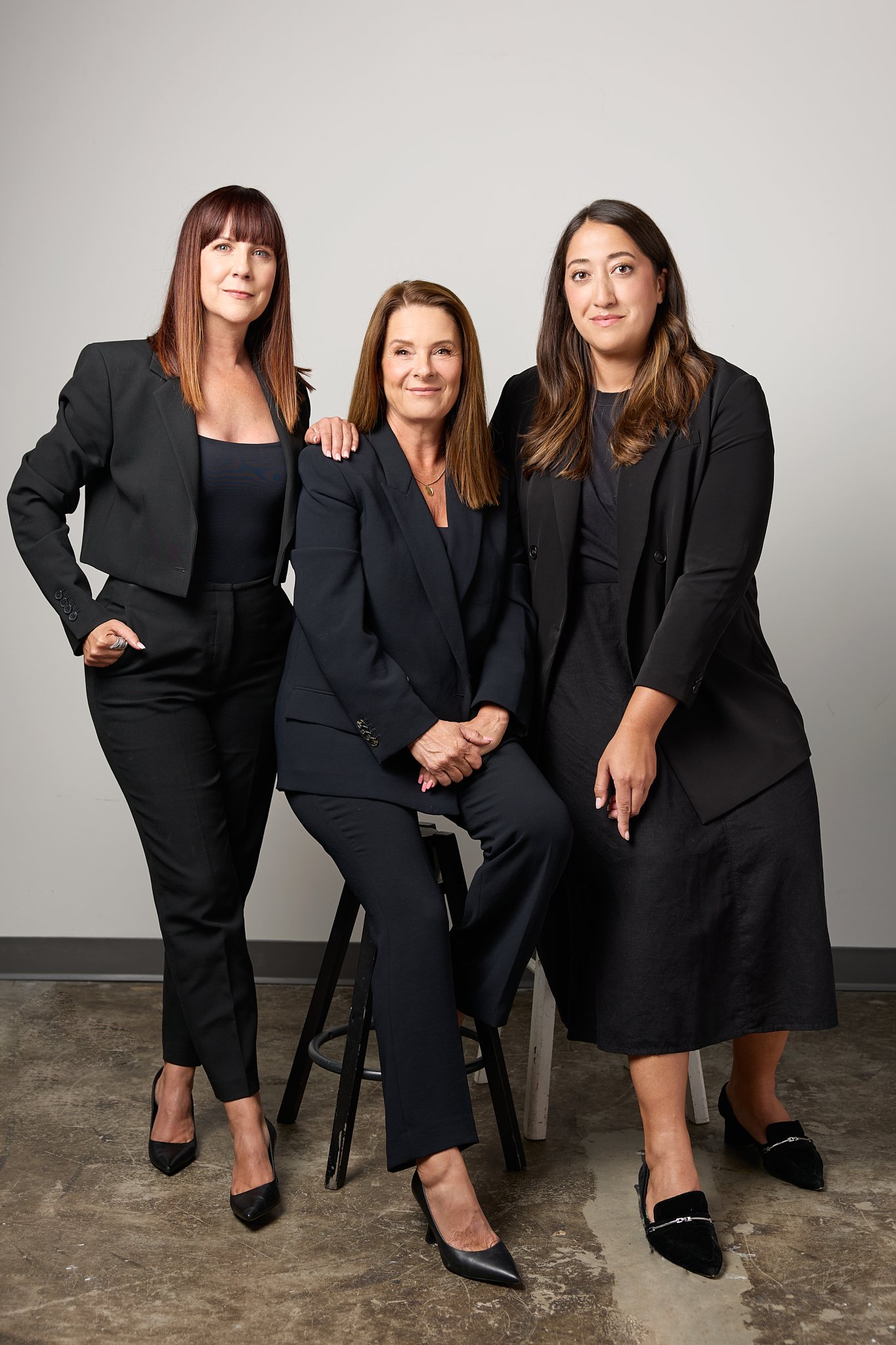 Three women in black business attire posing together indoors against a plain wall, two sitting on stools and one standing.