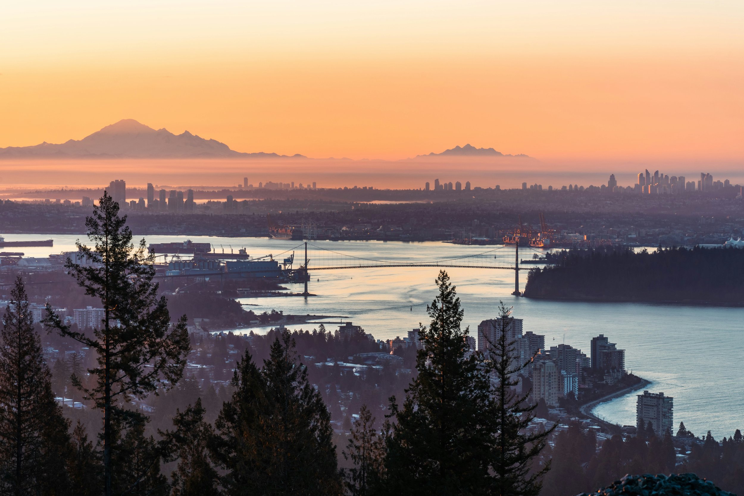 A city skyline overlooking water, with mountains in the background at sunset, and trees in the foreground.